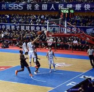 A basketball game takes place in a large indoor arena filled with spectators. Players from two teams, one in black uniforms and the other in white, compete for the ball near the hoop. The stadium is adorned with banners in Chinese, and a scoreboard displays a tied score of 21. The court features a distinctive blue and wood pattern.