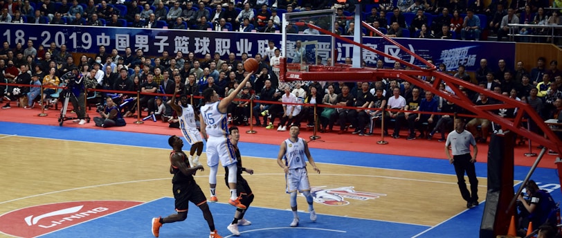 A basketball game takes place in a large indoor arena filled with spectators. Players from two teams, one in black uniforms and the other in white, compete for the ball near the hoop. The stadium is adorned with banners in Chinese, and a scoreboard displays a tied score of 21. The court features a distinctive blue and wood pattern.