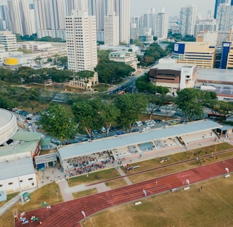 An aerial view of an urban area featuring a sports complex with a track field and a grandstand. Surrounding the sports area are numerous high-rise buildings, greenery, and roads with vehicles. The image captures people engaging in activities on the field.