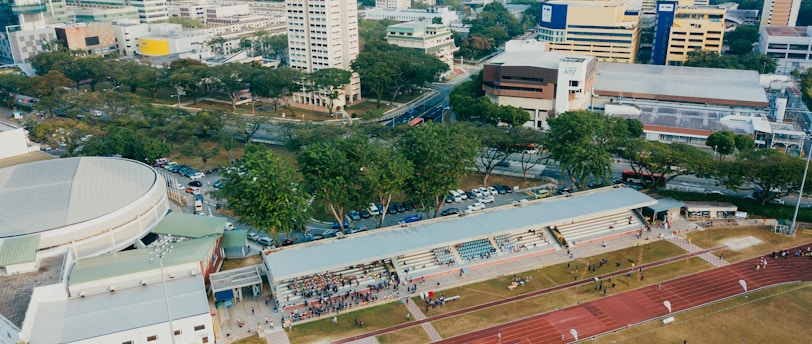 An aerial view of an urban area featuring a sports complex with a track field and a grandstand. Surrounding the sports area are numerous high-rise buildings, greenery, and roads with vehicles. The image captures people engaging in activities on the field.