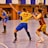 A dynamic indoor sports scene with players in motion on a court. Two athletes in blue and yellow uniforms are engaged in an intense handball game, with one player gripping the other's jersey. The background shows other players, several seated people, and advertisements on the walls.