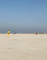 A group of people wearing colorful sports uniforms are playing cricket on a wide, open sandy ground. The sky is clear with a subtle gradient from blue to grey on the horizon. In the distance, there are structures that suggest a cityscape.