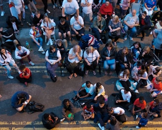 A large group of people gathered outdoors, watching a screen that is displaying a man. The crowd is diverse in age and appears interested and engaged, with many sitting closely together, some standing. Many are dressed casually, some in sports attire, and a few waving or holding flags. The event seems to have a communal, festive atmosphere.
