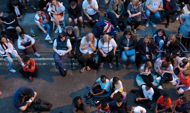 A large group of people gathered outdoors, watching a screen that is displaying a man. The crowd is diverse in age and appears interested and engaged, with many sitting closely together, some standing. Many are dressed casually, some in sports attire, and a few waving or holding flags. The event seems to have a communal, festive atmosphere.