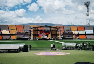 A sports stadium with a large stage set up on the field, hosting an outdoor event. Spectators wearing colorful hats sit in the foreground. The seating areas display vibrant colors, and a clear blue sky with scattered clouds is visible. Large screens display graphics related to the event.