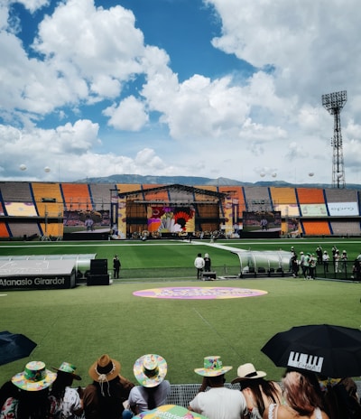 A sports stadium with a large stage set up on the field, hosting an outdoor event. Spectators wearing colorful hats sit in the foreground. The seating areas display vibrant colors, and a clear blue sky with scattered clouds is visible. Large screens display graphics related to the event.