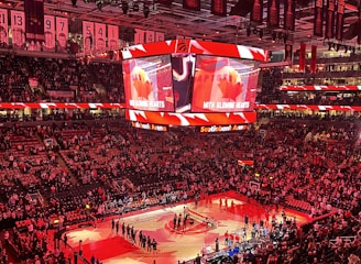 An indoor sports arena filled with spectators and a basketball court illuminated by red lighting. The scoreboards and video screens display promotional content, with numerous banners and championship flags hanging from the ceiling. Players and officials are gathered on the court, possibly in preparation for a game or event.