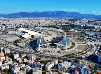 A large, modern sports complex is situated within an expansive urban landscape. The main stadium features prominent architectural structures and an open roof design. Surrounding the stadium are various sports facilities, roads, and greenery. Residential and commercial buildings extend into the distance, framed by mountain ranges under a clear blue sky.