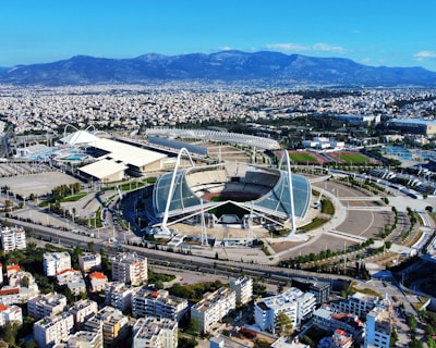 A large, modern sports complex is situated within an expansive urban landscape. The main stadium features prominent architectural structures and an open roof design. Surrounding the stadium are various sports facilities, roads, and greenery. Residential and commercial buildings extend into the distance, framed by mountain ranges under a clear blue sky.