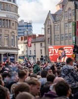A large crowd of people, many wearing red and white, gather in a city square. They are watching an event broadcast on a large screen, which displays a man speaking into a microphone with logos suggesting a sporting event. Surrounding buildings are historical and commercial, with banners indicating a connection to the Premier League.