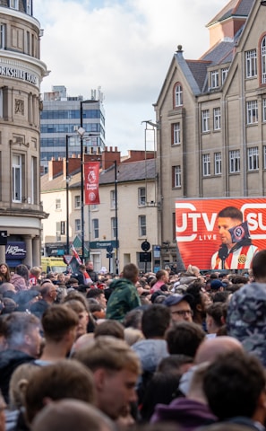 A large crowd of people, many wearing red and white, gather in a city square. They are watching an event broadcast on a large screen, which displays a man speaking into a microphone with logos suggesting a sporting event. Surrounding buildings are historical and commercial, with banners indicating a connection to the Premier League.