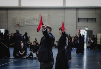 A martial arts event taking place in an indoor sports hall. Several individuals dressed in traditional martial arts attire participate, some wearing protective gear. A person prominently holds up a red flag, potentially indicating a referee or judge in action. The background includes spectators and sports equipment.
