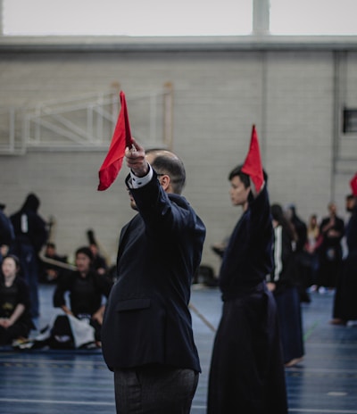 A martial arts event taking place in an indoor sports hall. Several individuals dressed in traditional martial arts attire participate, some wearing protective gear. A person prominently holds up a red flag, potentially indicating a referee or judge in action. The background includes spectators and sports equipment.