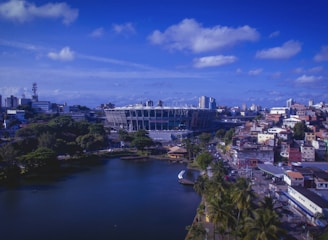 A vibrant cityscape with a large sports stadium at the center. The area around the stadium features a mix of modern and traditional buildings, with dense foliage and a peaceful body of water in the foreground. The sky is clear with scattered clouds, giving a bright and lively atmosphere.