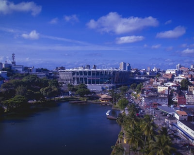 A vibrant cityscape with a large sports stadium at the center. The area around the stadium features a mix of modern and traditional buildings, with dense foliage and a peaceful body of water in the foreground. The sky is clear with scattered clouds, giving a bright and lively atmosphere.