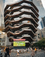 A large, spiral architectural structure with multiple levels stands prominently in an urban setting. In front of the structure, a large screen displays a sports broadcast, and several people are gathered around, some sitting and others standing. The scene is set outdoors, with towering modern buildings surrounding the area. The ground is paved with a geometric pattern of gray and white tiles.