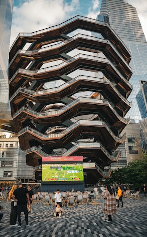 A large, spiral architectural structure with multiple levels stands prominently in an urban setting. In front of the structure, a large screen displays a sports broadcast, and several people are gathered around, some sitting and others standing. The scene is set outdoors, with towering modern buildings surrounding the area. The ground is paved with a geometric pattern of gray and white tiles.