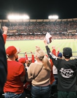 A large crowd of spectators is in a stadium during a sporting event, with many people raising their arms in excitement and wearing predominantly red clothing. The field is brightly lit and visible in the background, indicating an active scene.