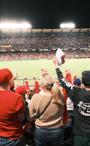A large crowd of spectators is in a stadium during a sporting event, with many people raising their arms in excitement and wearing predominantly red clothing. The field is brightly lit and visible in the background, indicating an active scene.